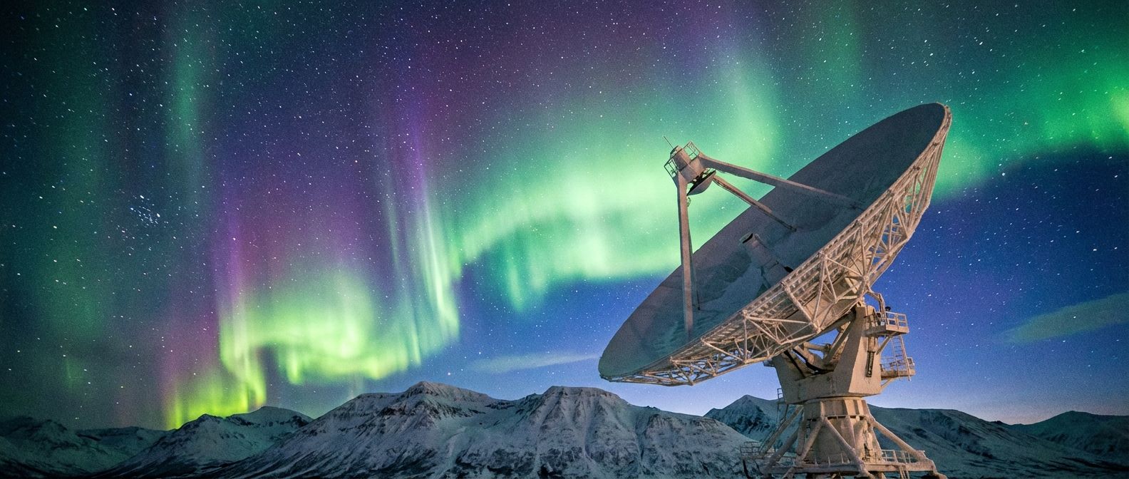 A dramatic wide-angle photograph of a vibrant aurora borealis dancing across a dark, star-filled night sky over a snow-covered mountain range. In the foreground, a large, elegant radio telescope dish points towards the celestial display, bathed in the ethereal light. The scene captures the awe-inspiring spectrum of natural and technological energy.
