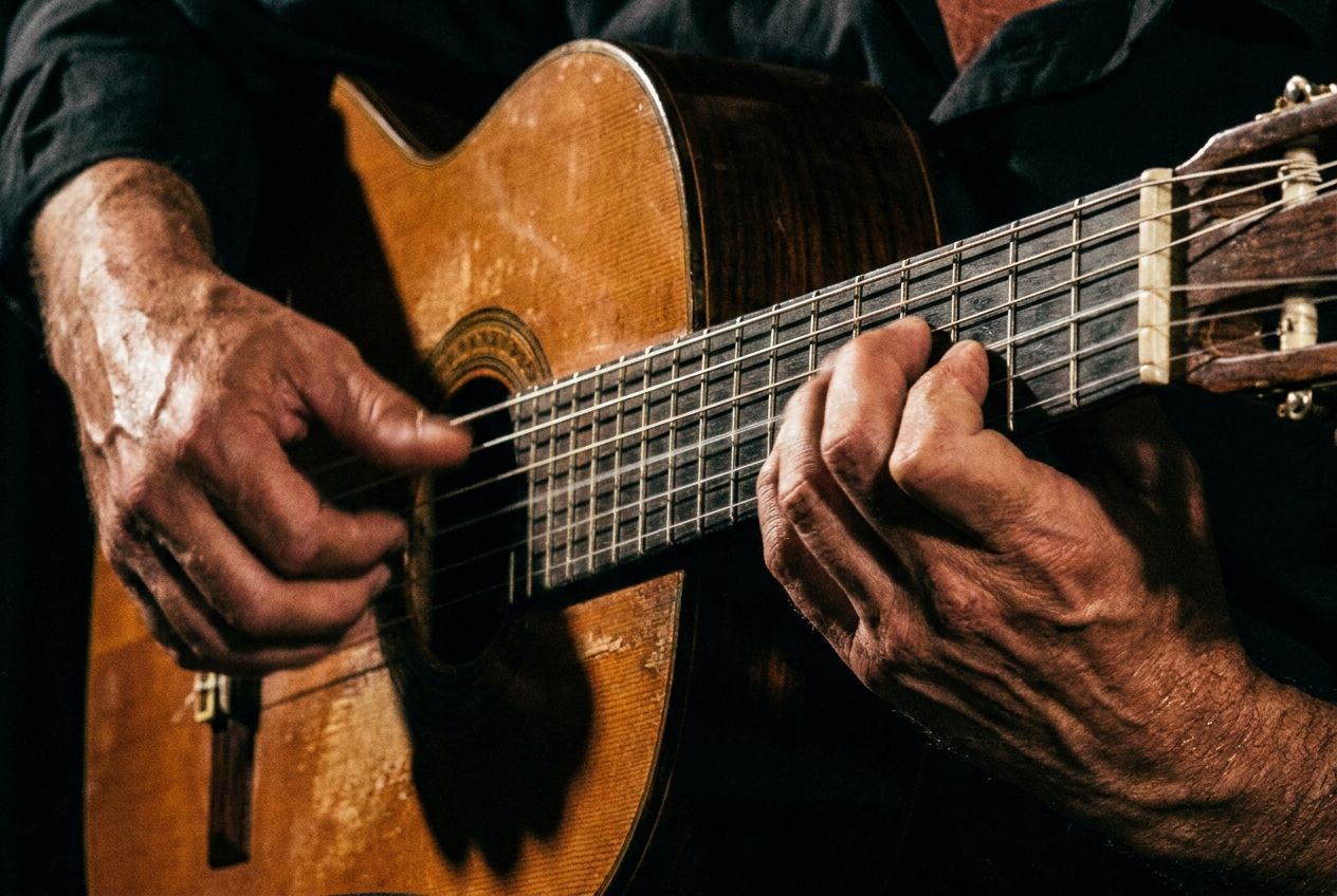 Una fotografía dramática y en primer plano de las manos de una persona tocando una guitarra clásica con gran destreza, los dedos moviéndose con agilidad sobre el diapasón y las cuerdas. La iluminación es intensa y resalta la habilidad y concentración del músico, capturando el momento de la ejecución con un encuadre horizontal 3:2.