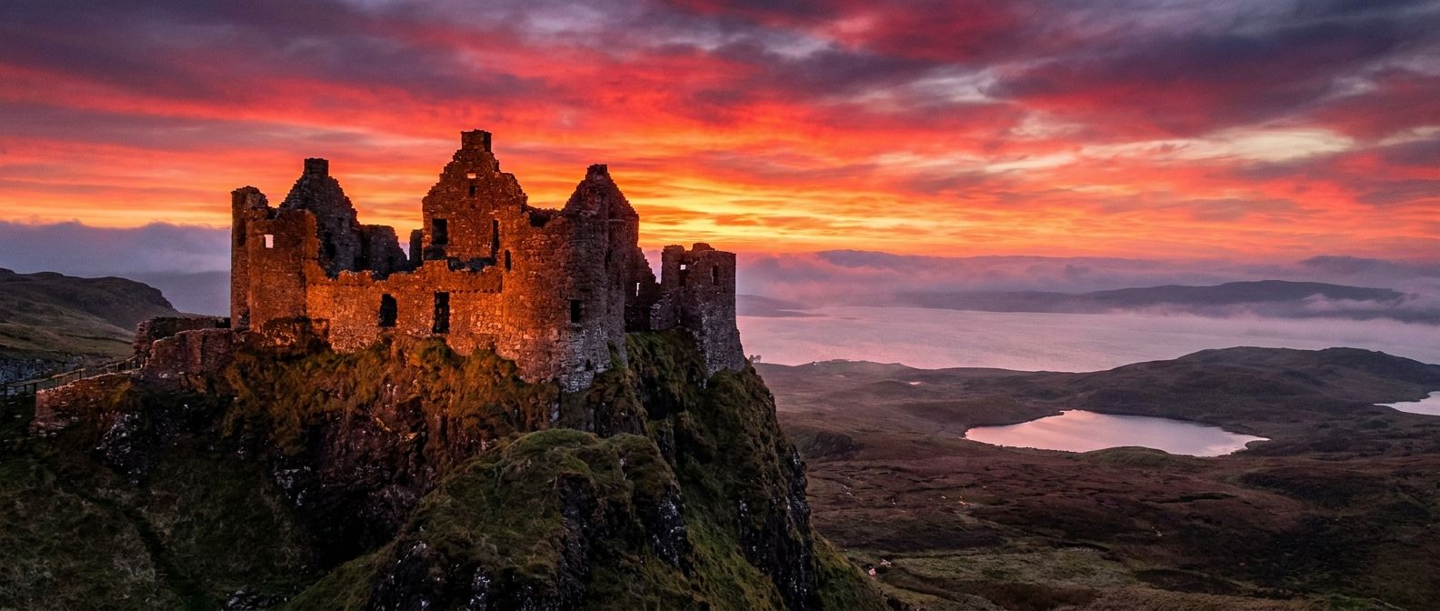 A dramatic wide-angle photograph of a majestic ruined medieval castle silhouetted against a vibrant sunset sky, perched on a rugged cliff overlooking a vast, misty Scottish loch. The ancient stone walls are illuminated by the warm, golden light, emphasizing the breathtaking scenery and rich British heritage.