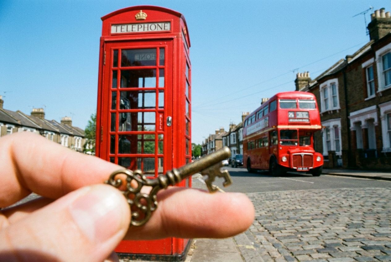 A dramatic, low-angle photograph captured in a 3:2 landscape frame. An extreme close-up of a hand holding a single ornate antique key in the foreground, with a shallow depth of field. In the mid-ground, a vibrant red British telephone box stands sharply in focus, while in the far distance, a classic red double-decker bus drives past a row of charming terraced houses under a bright, clear sky.