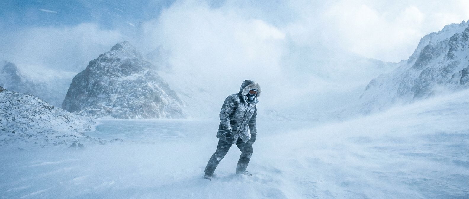 A solitary figure, bundled in heavy winter gear, struggles against a powerful blizzard in a dramatic, snow-swept landscape. Heavy snow swirls around them, emphasizing the absolute extreme cold, captured in a striking wide-angle photograph.