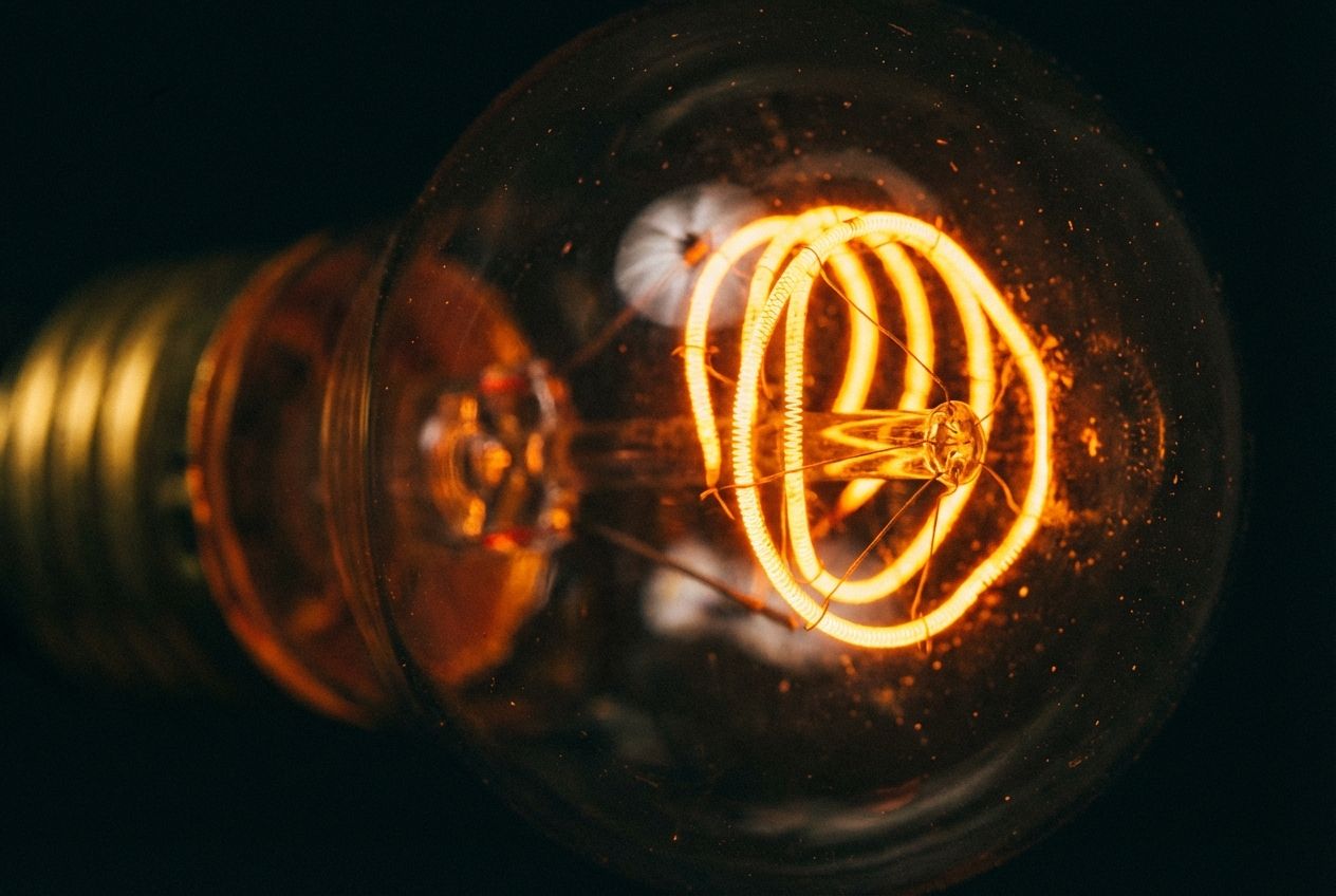 A dramatic, extreme close-up photograph of the glowing, incandescent filament inside a vintage light bulb. The coiled wire filament radiates a warm, bright orange-yellow light, contrasting with a dark, slightly blurred background, emphasizing the intense heat and light within the delicate structure, composed for a 3:2 landscape frame.