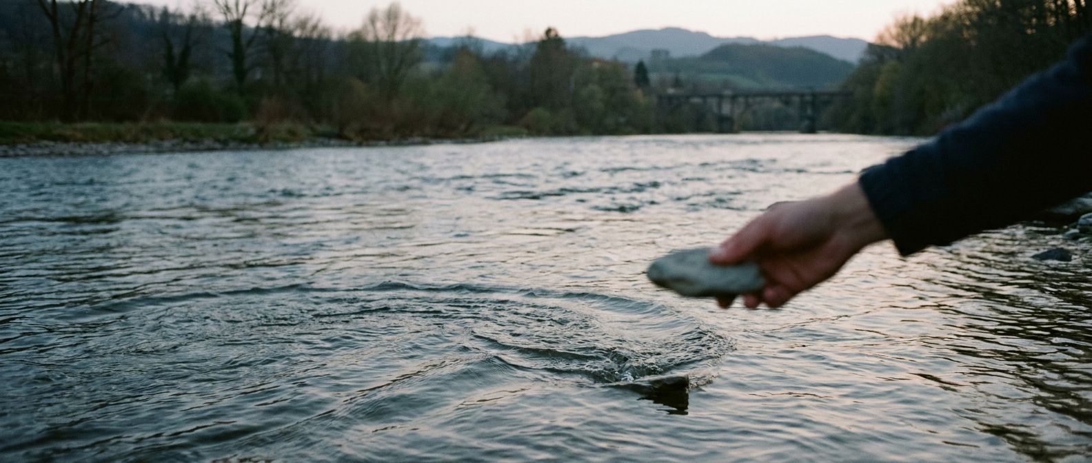 A wide, horizontal photograph captures a hand in motion, skipping a flat, grey stone across the calm surface of a wide, continuously flowing river. The stone creates a sharp, expanding ripple at the point of impact, contrasting with the river's unbroken current, all set against a tranquil, muted landscape under soft, ambient light at dusk.