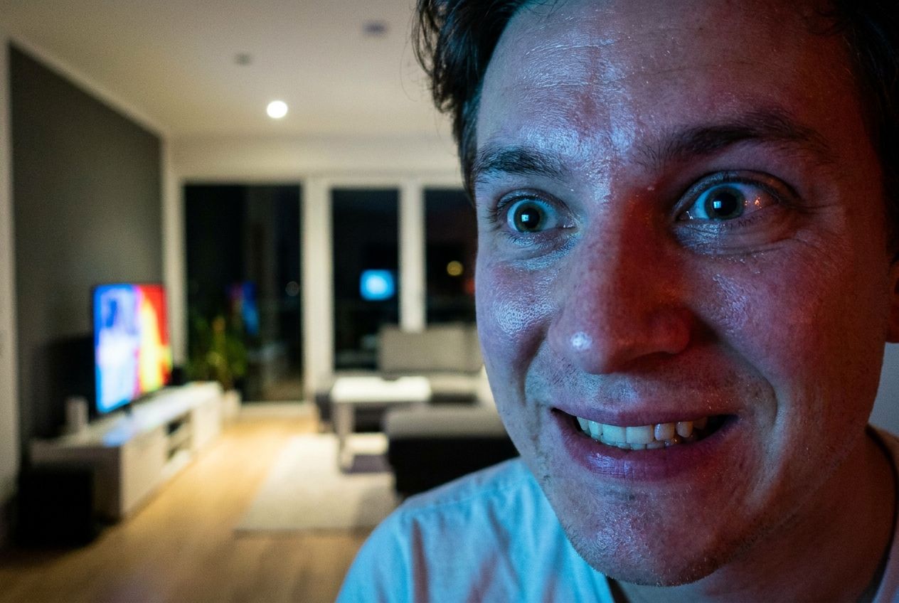 A dramatic close-up photograph of a person's face, illuminated by the dynamic, colorful light from a television screen, their expression one of intense excitement and suspense while watching a thrilling film or series. The background is a softly blurred, modern living room in a stylish German apartment, composed for a 3:2 landscape frame.
