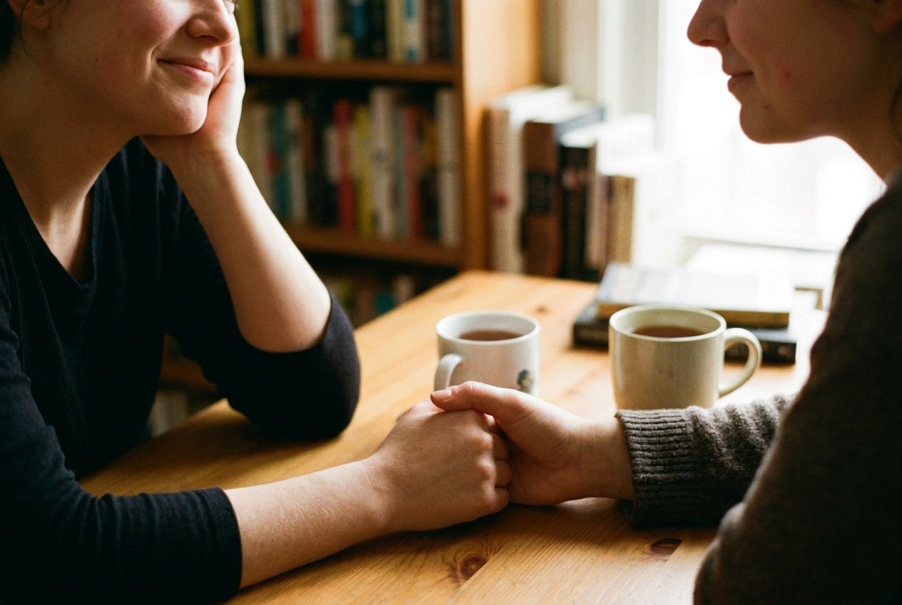 A close-up photograph of two individuals, one gently listening with an empathetic expression while the other speaks, their hands subtly clasped or gesturing, conveying a moment of shared understanding and advice in a warm, natural light. The setting is inviting and calm, composed for a 3:2 landscape frame.
