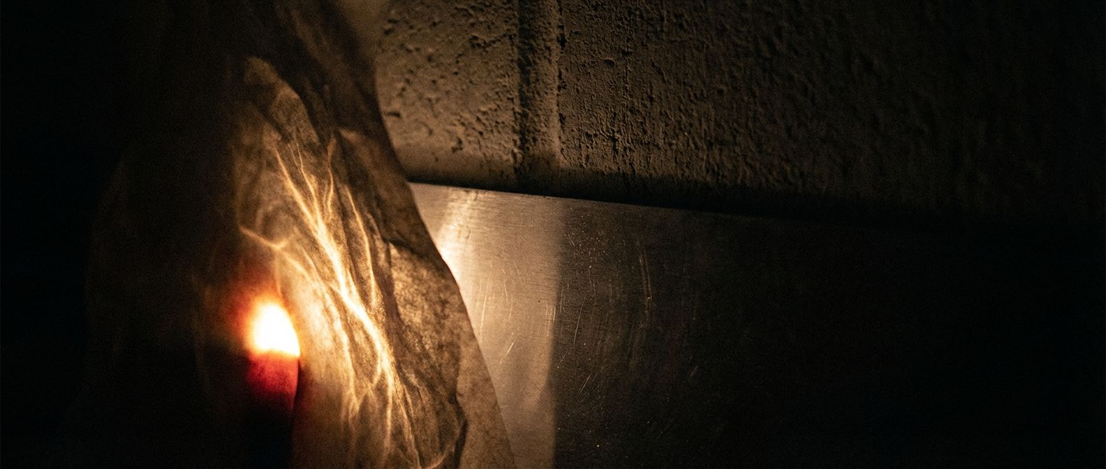 A dramatic, close-up photograph of an intense, ethereal glow emanating from a dark, unseen source. The powerful light pierces effortlessly through a delicate, crumpled sheet of paper, then struggles to faintly illuminate a thin sheet of brushed aluminum, before being completely absorbed by a thick, rough concrete wall in the background. The scene is cast in deep shadows with the glowing light as the sole, mysterious illumination, emphasizing the varying penetration of unseen forces in a wide, horizontal frame.