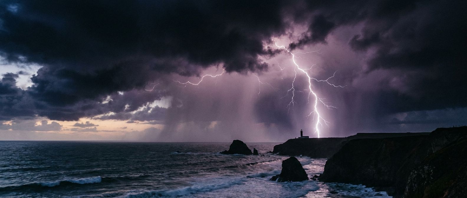 A wide, dramatic photograph of a powerful thunderstorm over a rugged coastline at dusk, with a jagged lightning bolt illuminating the dark, turbulent clouds and heavy rain. The scene should convey the raw, awe-inspiring force of a brewing gale, captured with intense mood.