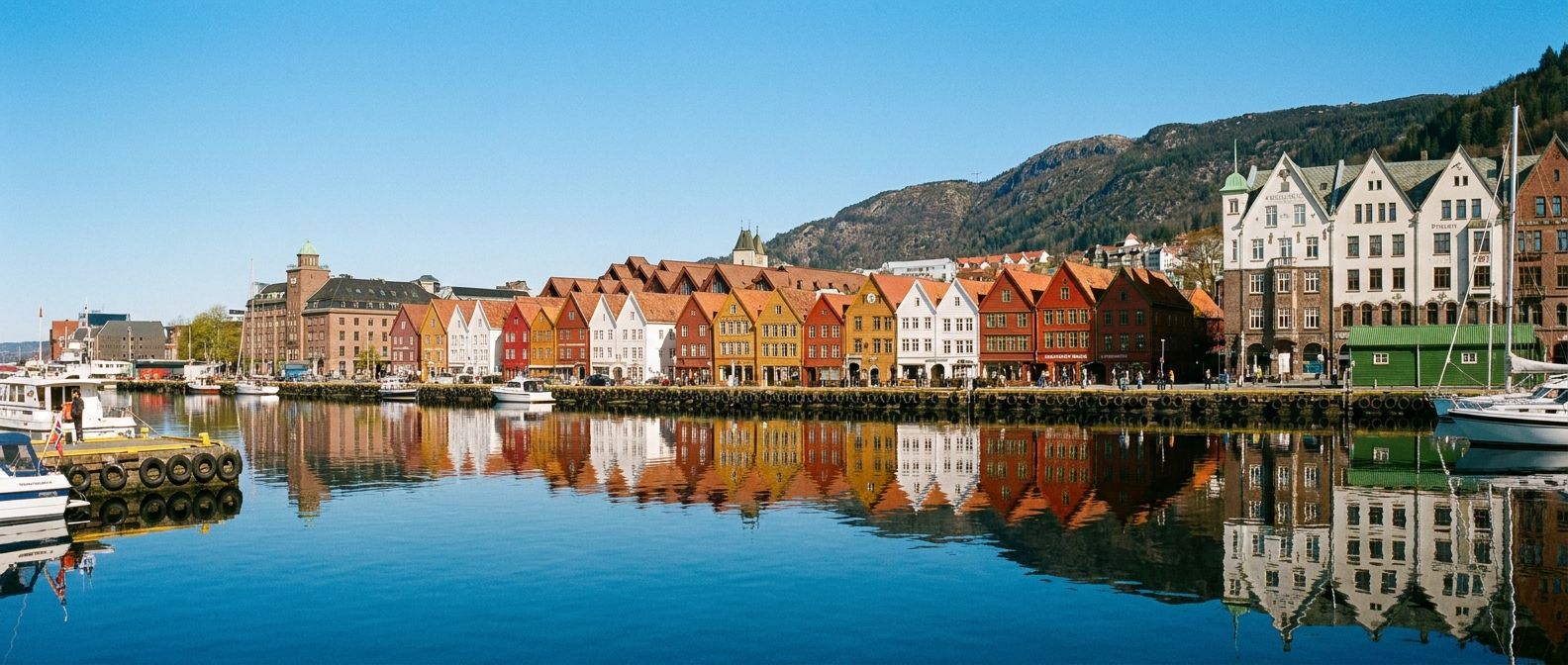 A vivid, wide-angle photograph of the iconic, colorful wooden Bryggen warehouses in Bergen, Norway, reflecting in the calm harbor waters. The vibrant red, yellow, and white facades stand out vividly under a clear, bright sky, showcasing the unique cultural charm of the city.
