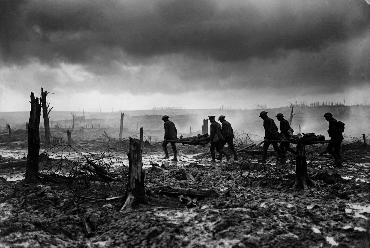 A dramatic wide-angle photograph of British stretcher bearers, silhouetted against a murky, shell-blasted landscape of the Western Front, struggling through deep mud and tangled barbed wire under a heavy, overcast sky. The scene emphasizes the desolation and extreme difficulty of evacuation in a 3:2 landscape frame.
