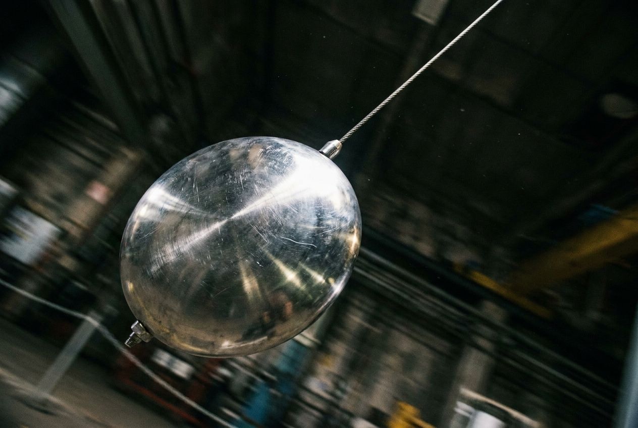 A dramatic, low-angle photograph of a large, heavy, polished steel pendulum at the very peak of its swing, momentarily motionless against a dark, blurred background. The scene is sharply focused on the pendulum, capturing the tension of potential energy and the precise moment before its inevitable descent, symbolizing a future event contingent on a present condition. The composition is for a 3:2 landscape frame, emphasizing the dramatic pause.