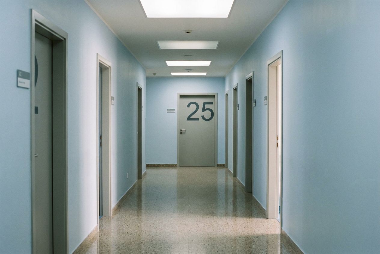 A long, empty corridor inside a clean, well-maintained medical polyclinic, with doors lining both sides. One door in the mid-distance prominently displays the number '25'. The scene is lit by soft, diffused overhead lighting, creating subtle shadows and a quiet, expectant atmosphere, composed for a 3:2 landscape photograph.