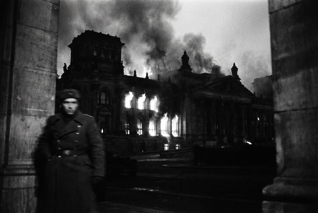 A black and white photograph of a bonfire in the Reichstag building, smoke billowing from the windows, with a blurred silhouette of a Nazi figure watching from the shadows. The image is high contrast and grainy, evoking a sense of chaos and foreboding.