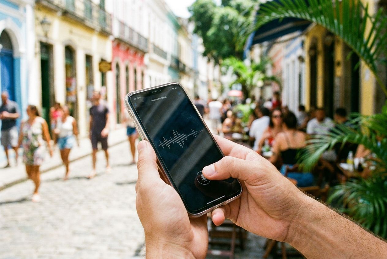 A close-up, dramatic photograph of a person's hands holding a modern smartphone, with a thumb hovering over the screen as if recording a voice message. The background is a vibrant, sun-drenched, blurred scene of a bustling Brazilian street or cafe, capturing the essence of daily life and connectivity in a 3:2 landscape frame with natural, bright lighting.